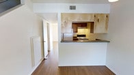 This two-floor residence offers a functional and welcoming layout designed for comfortable everyday living. The main living area features warm hardwood-style flooring that extends throughout, creating a cohesive and inviting atmosphere. A defining feature of the space is the open pass-through kitchen, which connects seamlessly to the living area and enhances the sense of openness. The kitchen is equipped with essential appliances, including a refrigerator and a range, and is framed by warm wood-toned cabinetry paired with a distinctive vertical shiplap accent wall that adds a touch of rustic character. The countertop bar ledge serves as a natural dividing point between the kitchen and living space, ideal for casual dining or entertaining. A hallway leads to additional rooms, suggesting a well-organized floor plan with clear separation between living and private spaces. Overhead, a textured ceiling and a centrally placed ventilation register point to a thoughtfully maintained interior. The neutral white walls and trim throughout allow natural light to reflect freely, keeping the space feeling bright and airy. Spanning two floors, this residence offers versatile living arrangements suited to a variety of needs.