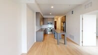 This well-appointed one-bedroom, one-bathroom unit offers a compact yet efficient layout across a single floor, with every square foot purposefully utilized. Light hardwood flooring runs throughout, lending warmth and visual continuity to the space.

The kitchen is a standout feature, showcasing contemporary gray cabinetry that extends to the ceiling for maximum storage, complemented by open wood-accented display shelving. A white subway tile backsplash, light quartz countertops, and a full suite of stainless steel appliances — including a range, over-the-range microwave, dishwasher, and refrigerator — complete the polished look. A central island with a built-in table extension provides both additional prep space and casual dining functionality.

A hallway connects the main living areas to the private bedroom, which features clean white walls and a tall window offering views of the surrounding urban streetscape, filling the room with natural light. A dedicated closet provides organized storage.

The bathroom includes a glass-enclosed shower with chrome fixtures, maintaining the unit's clean, modern aesthetic. The hallway also houses a spacious closet fitted with a stacked washer and dryer, adding notable in-unit convenience. Recessed lighting and a neutral color palette throughout create a bright, cohesive atmosphere.