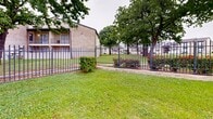 Located at 745 E Pecan St in Hurst, Texas, this single-level community amenity space centers around a generously sized oval swimming pool surrounded by a wide concrete deck, providing ample room for lounging and recreation. The pool area is enclosed by a sturdy wrought iron fence, ensuring safety and security for residents, and is complemented by well-maintained green lawns and mature shade trees that add a welcoming, park-like atmosphere.

Adjacent to the pool, a brick-and-navy-accented clubhouse building features multiple glass-panel doors and covered entryways supported by brick columns, offering a functional and inviting gathering space for the community. The building's design blends classic brick construction with bold navy accent colors for a clean, contemporary look.

The surrounding grounds are thoughtfully landscaped with trimmed shrubs, flowering plants, and expansive grassy areas framed by mature oak trees, creating a pleasant outdoor environment. The perimeter iron fencing with gated access contributes to the community's secure and organized layout. Standard pool safety signage is posted, noting no lifeguard on duty and no diving. The overall setting presents a well-maintained, community-oriented outdoor space suited for recreation and relaxation in a residential neighborhood.