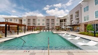 This outdoor pool area in Gonzales, Louisiana, presents a well-appointed resort-style amenity space designed for relaxation and recreation. The centerpiece is a generously sized swimming pool with clear, blue-tinted water, featuring a shallow tanning ledge along one side where lounge chairs invite sunbathing directly at the water's edge. Pool depth markers are visible, reflecting attention to safety throughout the space.

The expansive pool deck is finished in light-toned concrete, providing ample room for circulation and leisure. Multiple seating arrangements are distributed across the deck, including lounge chairs, dining-height tables, and chairs shaded by teal market umbrellas. A wooden pergola structure anchors one side of the deck, offering a covered gathering spot and adding architectural warmth to the otherwise contemporary surroundings.

Carefully placed landscaping with ornamental grasses and mulched planting beds introduces natural texture and visual contrast against the hard surfaces. The surrounding multi-story building features a modern exterior combining board-and-batten siding with brick accent walls—an exposed brick detail also visible in close-up, showcasing a variegated, light-toned masonry finish. Glass entry doors with clean metal hardware provide access between the pool courtyard and interior spaces, completing this well-designed outdoor amenity area.