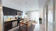 Located at 15741 SW 99th Pl in Miami, FL, this single-floor residence offers a thoughtfully designed open-concept layout that seamlessly connects the kitchen, dining, and living areas. The kitchen is outfitted with dark espresso cabinetry, granite countertops, a mirrored backsplash, and a full suite of stainless steel appliances, including a French door refrigerator, electric range, built-in microwave, and dishwasher. A pendant light fixture anchors the dining area, where a warm wood dining table provides an elegant space for entertaining. The living area is bright and airy, featuring a large floor-to-ceiling window dressed with sheer white curtains that flood the room with natural light. A full-wall mirror expands the sense of space and reflects the room's clean, contemporary aesthetic. Light wood flooring runs throughout, complementing the neutral color palette. The bedroom is a serene retreat, offering ample natural light through sheer-curtained windows, mirror panel accents, and a wall-mounted media setup. The bathroom features a floating dark-wood vanity with a granite countertop, an undermount sink, and a well-lit mirror, maintaining the cohesive modern design found throughout the residence.