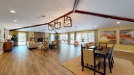 Located at 1300 Southampton Rd in Benicia, California, this single-floor community clubhouse offers a thoughtfully designed gathering space that blends warmth and functionality. The centerpiece of the space is a grand open great room with soaring vaulted ceilings accented by rich dark wood beams and striking geometric pendant chandeliers. A full-wall stacked stone fireplace anchors one end of the room, creating a cozy focal point surrounded by multiple comfortable seating arrangements. Warm hardwood-style flooring flows seamlessly throughout the entire space, adding continuity and a welcoming feel. An expansive wall of multi-pane glass doors and windows floods the great room with natural light and provides direct views and access to an outdoor patio area. A dedicated dining area features a large dark wood table and is complemented by an adjacent console with a flat-screen display. The modern kitchen is outfitted with white shaker cabinetry, dark granite countertops, stainless steel appliances, a mosaic tile backsplash, and a glass door leading to the outdoors. A separate game room with a billiard table rounds out the amenities. Throughout, the warm color palette of yellows, oranges, and neutrals contributes to an inviting and vibrant atmosphere well-suited for community gatherings.