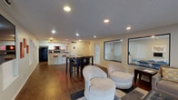 This well-appointed single-floor space is designed to serve as a versatile community or amenity hub, combining social, functional, and collaborative areas in one cohesive layout. The open-concept main area features rich hardwood flooring throughout, recessed lighting, and a comfortable lounge zone anchored by a large sectional sofa with accent pillows, complemented by a pair of plush swivel chairs and an oversized tufted leather ottoman — ideal for relaxed gathering or informal meetings. Large interior windows connect the lounge to adjoining rooms, creating a sense of openness and visual flow across the space. The kitchen is a standout feature, boasting dark granite countertops, white cabinetry, black appliances including a range and built-in microwave, and a double stainless steel sink. A curved stone-clad breakfast bar with bold pendant lighting and stylish red barstools adds a distinctive, modern flair. Adjacent to the kitchen, a bistro-style high-top table with bar chairs provides additional dining or work seating. The space also includes dedicated workstation areas and a glassed-in conference room with seating for small groups, making it highly functional for productivity. The overall ambiance is modern and inviting, with warm tones, decorative artwork, and thoughtful design details throughout.