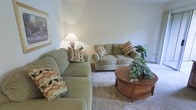 This single-floor residential space offers a warm and inviting atmosphere designed for comfortable everyday living. The living room serves as the heart of the home, featuring plush, sage-green upholstered sofas adorned with decorative throw pillows in earthy tones of brown, rust, and green. The seating arrangement is thoughtfully organized around a rich wooden oval coffee table, which adds a classic, grounded focal point to the room. A potted green plant atop the coffee table brings a touch of nature indoors, complementing the lush greenery placed nearby. Soft, wall-to-wall carpeting in a neutral tone enhances the cozy ambiance underfoot, while smooth, light-colored walls provide a clean and airy backdrop. A table lamp with a warm-toned shade casts a gentle glow, contributing to the relaxed and homey feel of the space. A framed piece of artwork adorns one wall, adding a subtle decorative accent. The overall design aesthetic is traditional and understated, prioritizing comfort and functionality. The space feels well-proportioned and organized, making it a practical and pleasant environment for relaxation and everyday use.