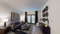 This single-floor residence is thoughtfully designed with a cohesive contemporary aesthetic carried throughout each space. The living room serves as the focal point, featuring wood-look flooring, a bold black-and-white patterned area rug, and a tufted charcoal sofa centered around a glass-top coffee table. A set of French doors with floor-to-ceiling dark drapes invites natural light while providing outdoor access. Decorative circular wall art adds visual interest alongside a large framed artwork piece.

The kitchen is outfitted with slate-gray cabinetry, granite countertops, and a full suite of stainless steel appliances, including a refrigerator, electric range with microwave above, and a deep sink. Light wood-look flooring continues from the entryway seamlessly into this space.

The bedroom offers a calm, neutral atmosphere with plush carpeting, natural light from a large window with blackout drapes, and ample room for a full furniture arrangement. The bathroom features granite countertops atop gray cabinetry, a wide vanity mirror, a glass-enclosed shower, and direct access to a walk-in closet with built-in shelving.