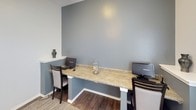 This single-floor space is designed as a functional shared workspace or business center. The room is finished with durable wood-look flooring and neutral-toned walls in soft gray and white, creating a clean, professional atmosphere. A built-in L-shaped workstation with a granite-style countertop anchors one side of the room, accommodating two computer workstations with monitors, keyboards, and accompanying seating. The countertop is supported by white pedestals and offers ample surface area for work tasks. A small decorative vase and glass jar add subtle finishing touches to the station.

The room benefits from multiple windows fitted with white horizontal blinds, allowing for controlled natural light. A glass-paneled entry door also contributes to the brightness of the space while maintaining visibility into adjacent areas. A fire extinguisher is mounted near the entry door, reflecting safety compliance. A woven area rug anchors the open central floor space, where additional seating, including a sleek black chair and a padded upholstered chair, provides flexible use of the room. The overall layout is open and uncluttered, supporting a productive and welcoming environment.