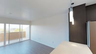 Located at 2799 Adrian in San Diego, this two-floor residence at Loma21 offers a contemporary living experience defined by clean lines and modern design. The living area is open and airy, featuring smooth white walls, high ceilings, and durable wood-look flooring that adds warmth to the minimalist aesthetic. A sliding glass door fills the space with natural light and provides direct access to a private balcony, offering a seamless connection to the outdoors. Stylish pendant lighting with a chrome finish adds a refined touch to the interior, complementing the rich dark wood accents seen on surrounding cabinetry and doors. The kitchen features light-toned countertops that contrast elegantly with the darker design elements, creating a balanced and modern palette throughout. The two-floor layout provides a sense of dimension and separation between living spaces, making for a functional and well-organized home. Overall, the unit presents a thoughtfully designed environment with a cohesive modern aesthetic, quality finishes, and practical features suited for comfortable urban living in San Diego.