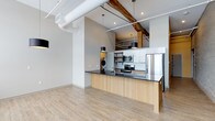 This single-floor loft seamlessly blends industrial character with contemporary design. Soaring ceilings draw the eye upward, where exposed wooden beams and visible ductwork pay homage to the building's raw architectural heritage, while modern finishes bring a polished, up-to-date aesthetic throughout. The open-concept layout allows the living area and kitchen to flow together effortlessly, creating a spacious and airy environment enhanced by light hardwood flooring that runs throughout the space. The kitchen serves as a central focal point, anchored by a generous island with a dark stone countertop and light wood cabinetry beneath. Stainless steel appliances, a subway tile backsplash, and open shelving complete the culinary space with both function and style. Pendant lighting in matte black hangs above the island, complementing the industrial tone set by the exposed overhead pipes and beams. A dedicated laundry area is conveniently tucked within the unit, adding practical everyday functionality. The overall ambiance is modern yet warm, with a thoughtful balance of raw structural elements and refined finishes that give this loft a distinctive, character-rich personality.