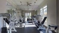 This fitness center is designed to offer a comprehensive workout experience within a bright and well-appointed space. The room features large windows along multiple walls, flooding the interior with an abundance of natural light and providing views of the surrounding outdoor environment. Ceiling fans mounted overhead ensure comfortable air circulation throughout the space during workouts.

The flooring consists of a durable dark rubberized surface, well-suited for heavy equipment use and providing traction and safety for users. Full-length mirrors line the walls, enhancing the sense of openness while allowing users to monitor their form during exercise.

The facility is outfitted with an extensive selection of equipment, including cable and weight machines for targeted strength training, a rowing machine, adjustable benches with free weights, and a variety of cardio machines such as ellipticals and stair climbers. The equipment is arranged in an organized layout that maximizes available floor space while maintaining clear movement paths.

Architectural details such as crown molding and a bay window area add a refined aesthetic to the space, elevating it beyond a purely utilitarian environment. The overall ambiance is clean, functional, and inviting, making it a well-rounded fitness facility suited for a broad range of workout routines.