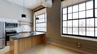This single-floor space masterfully combines industrial heritage with modern design, creating a distinctive and functional environment. Exposed brick walls serve as a defining architectural element, lending warmth and texture throughout the interior. Large, multi-pane windows with bold black steel frames flood the space with abundant natural light while framing views of the surrounding urban landscape.

The kitchen area is thoughtfully designed with warm wood cabinetry, a sleek dark countertop, and a undermount sink paired with a modern faucet. Open shelving mounted against a white subway tile backsplash provides both storage and display space, while pendant lighting with cylindrical black shades adds a refined industrial touch overhead.

The polished concrete flooring ties the space together, offering a durable yet visually appealing surface that complements the raw brick and natural wood tones. Exposed ductwork running along the upper portion of the walls reinforces the loft-inspired aesthetic, adding an authentic industrial character. The overall ambiance is one of curated contrast — where rugged materials meet clean, contemporary lines — making this a versatile and visually compelling space.