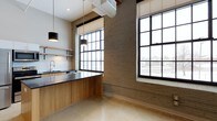 This single-floor space masterfully combines industrial heritage with modern design, creating a distinctive and functional environment. Exposed brick walls serve as a defining architectural element, lending warmth and texture throughout the interior. Large, multi-pane windows with bold black steel frames flood the space with abundant natural light while framing views of the surrounding urban landscape.

The kitchen area is thoughtfully designed with warm wood cabinetry, a sleek dark countertop, and a undermount sink paired with a modern faucet. Open shelving mounted against a white subway tile backsplash provides both storage and display space, while pendant lighting with cylindrical black shades adds a refined industrial touch overhead.

The polished concrete flooring ties the space together, offering a durable yet visually appealing surface that complements the raw brick and natural wood tones. Exposed ductwork running along the upper portion of the walls reinforces the loft-inspired aesthetic, adding an authentic industrial character. The overall ambiance is one of curated contrast — where rugged materials meet clean, contemporary lines — making this a versatile and visually compelling space.