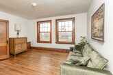 This single-floor space combines classic architectural details with functional, updated interiors. Throughout the unit, warm hardwood floors with rich tones create a cohesive and inviting atmosphere, complemented by white walls and dark wood trim that frames doorways and windows.

The living area is generously proportioned, accommodating a full-sized sofa with ease. Dual double-hung windows trimmed in dark wood allow natural light to filter in, offering views of the surrounding neighborhood. The open layout visible in the floor plan reveals a seamless flow between the living space and other areas of the unit.

The kitchen features white cabinetry with sleek brushed-metal hardware and a bold black countertop that provides strong visual contrast. An electric range and a single-basin sink are neatly integrated into the layout, offering practical functionality in a compact footprint.

The bathroom maintains a clean, bright aesthetic with white walls and classic white penny-tile flooring. A full tub-shower combination and a wall-mounted sink complete the space, keeping the design simple and timeless.

Overall, the space strikes a balance between the charm of older architectural elements—such as the substantial wood trim and hardwood floors—and modern, updated fixtures that enhance everyday livability.