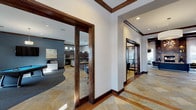 This single-story clubhouse is designed with a refined yet inviting aesthetic, blending upscale finishes with functional amenity spaces. The entry foyer sets the tone with large-format stone tile flooring featuring warm earth tones, dark wood door and window surrounds, decorative ironwork entry doors, and contemporary artwork displayed along crisp white walls. A circular pendant light fixture anchors the entryway ceiling.

The main lounge area is a standout, featuring soaring coffered ceilings painted in a deep teal-blue, flanked by rich dark wood beams, and a stone-clad fireplace with built-in display niches. Sheer curtains frame French doors that open to an outdoor area, allowing natural light to fill the space. Comfortable seating and curated décor create a welcoming atmosphere.

The game room provides a dedicated entertainment area complete with a pool table, shuffleboard table, wall-mounted television, and bar-height seating. The kitchen is fully equipped with dark granite countertops, rich cabinetry, a gas cooktop, and modern ring pendant lighting beneath high clerestory windows. A business center with workstations is also accessible. Throughout, consistent stone tile flooring, dark wood trim, and recessed lighting tie the spaces together cohesively.