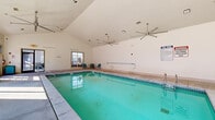 Located at 8064 S Fulton Pkwy in South Fulton, Georgia, this single-level indoor pool facility offers a clean and functional aquatic environment designed for year-round use. The centerpiece of the space is a generously sized rectangular swimming pool with clear turquoise water, tiled interior walls, and clearly marked depth indicators along the edges. Entry into the pool is made easy via stainless steel handrails and a gradual step entry at the shallow end.

The architecture of the room is highlighted by soaring vaulted ceilings that lend an open, airy feel to the space. Large ceiling fans are mounted overhead to assist with air circulation, maintaining a comfortable environment throughout. Expansive floor-to-ceiling windows and glass-paned doors line the perimeter, allowing abundant natural light to flood the interior and providing views of the surrounding outdoor greenery.

The pool deck, finished in a smooth concrete surface, provides ample space for movement around the pool. A small seating area with teal-colored chairs and a table is positioned near one of the windows, offering a relaxed spot for poolside lounging. Posted pool rules and safety signage are prominently displayed, and a life ring is mounted on the wall, underscoring the facility's commitment to safety. The overall ambiance is bright, functional, and well-maintained.