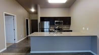 This single-floor space showcases a contemporary open-concept design centered around a well-appointed kitchen. The kitchen is equipped with a full suite of stainless steel appliances, including a refrigerator, electric range, and built-in microwave. Dark espresso-toned cabinetry lines the walls, providing ample storage, and contrasts cleanly against the neutral beige walls. A generous peninsula with a light-colored countertop and an integrated sink serves as both a functional workspace and a natural divider between the kitchen and the adjoining living area. Recessed and flush-mounted ceiling lighting ensures the kitchen is well-lit throughout. The flooring transitions from dark hardwood-style material in the kitchen zone to carpet in the living area, subtly defining each space. A hallway visible from the main area leads to additional rooms, suggesting a practical and organized floor plan. The overall aesthetic is clean and modern, with a neutral color palette punctuated by the dark cabinetry, lending the space a polished, move-in-ready appeal.