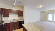 Located at 5721 Goodstone Dr in Raleigh, North Carolina, this single-floor residence offers a clean, functional layout designed for comfortable everyday living. The kitchen is a standout feature, showcasing rich dark-wood cabinetry with sleek hardware, light granite countertops with subtle veining, and a built-in sink positioned beneath recessed cabinetry. A kitchen island with a matching granite surface provides additional prep space and serves as a natural divider between the kitchen and the adjoining living area. Black appliances, including a dishwasher, complement the warm tones of the cabinetry. Track lighting above the kitchen ensures ample task illumination. The kitchen flows seamlessly into a spacious, open living area finished with neutral carpet flooring and warm white walls, creating a bright and airy ambiance throughout. The open sightlines between the kitchen and living space make the layout feel expansive and well-connected, ideal for both daily routines and entertaining. A doorway visible from the living area provides access to additional rooms, reinforcing the practical and efficient flow of the home. The overall design balances warmth and simplicity with quality finishes throughout.
