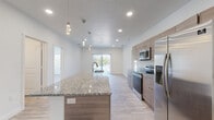 This contemporary single-floor residence is designed with a clean, modern aesthetic, showcasing light wood-style flooring and crisp white walls that create a bright, cohesive atmosphere throughout. At the heart of the home is an open-concept kitchen and living area, where a generously sized granite-topped island serves as a central focal point, complete with an integrated sink and pendant lighting above. The kitchen is fully equipped with stainless steel appliances — including a refrigerator, electric range, and microwave — alongside warm-toned cabinetry that adds a natural contrast to the light surroundings. Recessed lighting and access to a sliding glass door with vertical blinds allow natural light to fill the living space, which flows seamlessly from the kitchen. The bedrooms are comfortably sized, each featuring large windows with horizontal blinds that invite outdoor views and ample natural light. The primary bedroom includes a ceiling fan and notably large windows. Two bathrooms maintain the home's consistent design language, each featuring granite-topped vanities with wood-toned cabinetry and large mirrors. One bathroom includes a tub-shower combination, while the other features a walk-in shower. The overall ambiance of the space is clean, modern, and move-in ready.