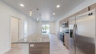 This contemporary single-floor residence is designed with a clean, modern aesthetic, showcasing light wood-style flooring and crisp white walls that create a bright, cohesive atmosphere throughout. At the heart of the home is an open-concept kitchen and living area, where a generously sized granite-topped island serves as a central focal point, complete with an integrated sink and pendant lighting above. The kitchen is fully equipped with stainless steel appliances — including a refrigerator, electric range, and microwave — alongside warm-toned cabinetry that adds a natural contrast to the light surroundings. Recessed lighting and access to a sliding glass door with vertical blinds allow natural light to fill the living space, which flows seamlessly from the kitchen. The bedrooms are comfortably sized, each featuring large windows with horizontal blinds that invite outdoor views and ample natural light. The primary bedroom includes a ceiling fan and notably large windows. Two bathrooms maintain the home's consistent design language, each featuring granite-topped vanities with wood-toned cabinetry and large mirrors. One bathroom includes a tub-shower combination, while the other features a walk-in shower. The overall ambiance of the space is clean, modern, and move-in ready.