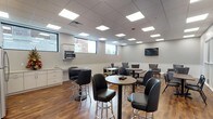 This well-appointed single-floor space is designed to serve as a comfortable and functional gathering area, blending casual dining, lounge, and collaborative zones seamlessly. The open layout is anchored by warm hardwood-style flooring that contrasts elegantly with the neutral gray walls and crisp white wainscoting that runs along the lower portion of each wall.

The main area is furnished with a mix of high-top bistro tables paired with sleek black leather bar stools, as well as standard-height wooden tables with matching black chairs, offering flexible seating for both quick gatherings and longer meetings. A dedicated lounge corner features plush gray upholstered armchairs arranged around a small round accent table, providing a relaxed setting beneath a wall-mounted flat-screen television.

The space is illuminated throughout by recessed LED panel lighting set within a drop-tile ceiling, creating a consistently bright and inviting atmosphere. Horizontal windows along the walls bring in natural light and offer views of the surrounding exterior brick architecture.

A built-in kitchenette area is equipped with white cabinetry, light stone-look countertops, a stainless steel microwave, a full-size refrigerator, and a sink — making the space fully self-sufficient for daily use. Glass-paneled interior walls and doors add transparency and an open feel to the overall design.
