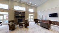 This single-floor common lounge area is characterized by its impressive architectural design, most notably the soaring coffered ceiling that amplifies the sense of openness throughout the space. Natural light streams in generously through a series of clerestory windows positioned high along the walls, brightening the neutral beige and warm taupe color palette that defines the interior.

The focal point of the room is a floor-to-ceiling stacked stone fireplace, complete with a classic mantel adorned with a traditional clock and lantern-style accents, providing a cozy and inviting atmosphere. Arranged around the fireplace is a thoughtfully curated seating area featuring patterned upholstered armchairs and a wooden coffee table, ideal for relaxed conversation.

A wainscoting detail along the lower portion of the walls adds a refined, traditional character to the space. On the opposite side of the room, additional seating is available alongside a sleek wooden credenza with decorative accessories. Glass-paneled doors at the rear of the space offer views of and access to an outdoor patio area, further enhancing the airy, connected feel of the lounge.