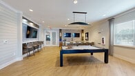 Located at 1851 Encantado Way in Kissimmee, Florida, this well-appointed community clubroom offers a modern and versatile gathering space designed for both recreation and relaxation. Light wood-look flooring flows throughout, complementing the crisp white walls and contemporary finishes that define the space's aesthetic.

At the heart of the main room sits a full-size pool table, illuminated by a sleek pendant light overhead. Adjacent to it, a large marble-topped island with bar-height seating provides an elegant surface for socializing or casual dining. A bold hexagonal-tile accent wall serves as a focal point along one side, flanked by a mounted flat-screen TV and warm sconce lighting. Additional bar-height counters with mounted displays are built into the shiplap-accented walls, offering flexible viewing and seating options.

Glass-paned French doors connect this recreation area to an adjoining lounge, which features soaring ceilings with exposed wood beams, a globe chandelier, and generous natural light from multiple windows. This open lounge area accommodates both dining-style seating and comfortable lounge chairs, making it suitable for a variety of gatherings. Throughout the space, carefully curated artwork and neutral décor contribute to a refined yet welcoming ambiance.