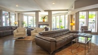 Located at 1001 Bolling Ave in Norfolk, Virginia, this single-floor community clubhouse offers a well-appointed and thoughtfully designed gathering space. The expansive main lounge area is anchored by rich wood-look flooring throughout and features multiple seating arrangements with contemporary leather and upholstered furniture. A sleek, built-in fireplace serves as a focal point in the lounge, complemented by interior windows that reflect natural light and enhance the sense of openness. Large exterior windows and glass doors flood the space with abundant natural light, creating a bright and welcoming ambiance. A dedicated coffee station with dark cabinetry, countertop appliances, and storage provides a convenient refreshment area. The full kitchen is outfitted with dark espresso cabinetry, granite countertops, a glass tile backsplash, and stainless steel appliances including a refrigerator, dishwasher, and microwave — ideal for hosting events or community gatherings. A separate business center offers workstations with computers and printers, along with comfortable seating, making it a functional space for residents to handle daily tasks. The overall design balances modern aesthetics with practical amenities, delivering a versatile and inviting community environment.