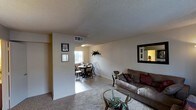 This two-floor residence features a thoughtfully arranged main level where the living and dining areas flow together in an open-concept layout. The living room is finished with wall-to-wall carpeting, neutral beige walls, and a textured ceiling, creating a warm and comfortable atmosphere. A large mirror with a dark frame adorns one wall, adding depth and reflected light to the space. Framed artwork provides subtle decorative touches throughout.

Adjacent to the living area, the dining space transitions to tile flooring and accommodates a round dining table with classic Windsor-style chairs. A window at the far end of the dining area invites natural light into the space, brightening the open floor plan. Overhead lighting provides additional illumination in the dining zone. Visible HVAC venting integrated into the ceiling ensures climate control throughout. The overall palette remains consistent with light, neutral tones across both areas, contributing to a cohesive and understated aesthetic. The combination of carpet and tile flooring clearly delineates the functional zones while maintaining a unified, approachable design throughout the main living level.