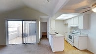This single-floor residence offers a bright and functional open-concept layout that seamlessly connects the kitchen and living areas. The kitchen is well-appointed with white cabinetry, a built-in range, an under-cabinet range hood, and a skylight above that floods the space with natural light. A central kitchen island with a granite-style countertop and integrated sink serves as both a practical workspace and a natural dividing point between the kitchen and the adjoining living area. The living space features plush carpeting and vaulted ceilings accented by an exposed beam, lending a sense of openness and architectural character. A textured ceiling adds a classic touch throughout. A large sliding glass door fitted with vertical blinds opens to an outdoor patio area, providing easy indoor-outdoor access and additional natural light. The transition from the carpeted living zone to the hardwood-style kitchen flooring creates a clear and visually appealing delineation between spaces. A hallway visible from the main living area leads to additional rooms, suggesting a thoughtful and practical floor plan designed for comfortable everyday living.