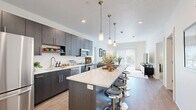 This thoughtfully laid-out single-story residence offers a comfortable and functional living experience across a well-utilized floor plan. The open-concept kitchen serves as a central highlight, featuring dark cabinetry with sleek hardware, stainless steel appliances including a refrigerator, microwave, and range, and a generous white-topped island with seating for multiple bar stools. Three pendant lights in brass tones hang above the island, adding a modern, stylish touch. Light wood-style flooring flows through the kitchen and into the adjoining living room, which benefits from large sliding glass doors that invite abundant natural light and outdoor views.

The living room is a bright, relaxed space with clean white walls and easy access to what appears to be an outdoor area. A dedicated office provides a quiet, versatile room suitable for work or study. The bedroom is a cozy retreat with carpeted flooring, a natural light-filled window, and direct access to one of the two bathrooms, which features a dark vanity with a white countertop, a tub-shower combination, wood-style flooring, and a walk-in closet. A second full bathroom with a wide vanity mirror serves guests. Two closets provide ample storage throughout.
