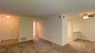 This single-floor space presents a straightforward, functional layout characterized by neutral cream-toned walls and consistent wall-to-wall carpeting throughout. The main living area is open and simply finished, with standard ceiling heights and a clean, understated aesthetic that provides a blank canvas for personalization.

The space features several HVAC vents, including both ceiling and floor-level units, ensuring climate control throughout. Electrical outlets are distributed along the baseboards at regular intervals. A centrally positioned wall divides the main living area, creating a subtle separation between spaces while maintaining an open sightline to adjoining rooms.

Visible from the main area, a hallway leads toward what appears to be additional rooms, including a well-lit corridor suggesting access to further living spaces. To the right, an adjacent area with its own carpeting extends the overall footprint of the unit, potentially serving as a dining area or secondary living space.

The overall ambiance is neutral and move-in ready, with consistent light-toned finishes and practical, no-frills details that prioritize functionality. The space benefits from interior lighting that keeps each area well-illuminated.