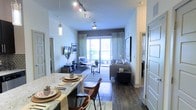 This single-floor residence showcases an open-concept design that seamlessly connects the dining and living areas into one cohesive, flowing space. Dark hardwood flooring runs throughout, providing a warm contrast against the light-toned walls and contributing to an inviting ambiance. The dining area is positioned near the kitchen and features a granite-topped counter with pendant lighting hanging above, adding a stylish focal point. Leather-accented chairs and thoughtfully arranged place settings complete the dining setup. The living area is generously proportioned, anchored by a comfortable sectional sofa, a glass-top coffee table, and a patterned area rug. A wall-mounted television and decorative artwork add functional and aesthetic character to the space. Track lighting runs along the ceiling, supplemented by a ceiling fan that enhances air circulation. Large sliding glass doors at the far end of the living room flood the interior with natural light and provide access to an outdoor area, framed by patterned draping curtains. The overall design blends modern and transitional elements, creating a bright, functional, and well-appointed living environment.