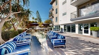 Located at 2420 Cardinal Drive in San Diego, CA, this outdoor courtyard space offers a well-appointed, resort-inspired amenity area designed for relaxation and social gathering. The grounds are finished with a sophisticated combination of patterned stone pavers and warm wood decking, including a distinctive circular deck inlay that serves as a central focal point for outdoor seating arrangements.

Generously sized blue-and-white striped lounge chairs and armchairs are arranged throughout the courtyard, providing ample seating in both shaded and sun-drenched areas. Large market umbrellas offer shade over additional dining and lounge areas, complementing the open-air atmosphere under San Diego's clear blue skies.

The landscaping is thoughtfully curated, featuring tall palms, sculptural dragon trees, ornamental grasses, and low-growing succulents housed in raised Corten steel planters that add a contemporary industrial touch. A linear water feature runs along one edge of the space, adding a tranquil visual element to the promenade-style walkway.

The modern multi-story building flanking the courtyard features clean white and grey facades, floor-to-ceiling glazing at ground level, and private balconies with sleek cable railings on upper floors. The overall ambiance is polished and inviting, blending Southern California's outdoor lifestyle with thoughtful architectural and landscape design.