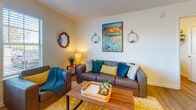 This thoughtfully designed 2-bedroom, 2-bathroom single-floor residence offers a comfortable and functional layout with consistent light wood-style flooring flowing throughout. The welcoming entryway, accented by a decorative wooden mirror and coat hooks, opens into a bright living room bathed in natural light from multiple large windows with slatted blinds. The living area is generously sized, providing ample room for a sectional sofa, accent chairs, and a coffee table, all complemented by warm, eclectic décor including landscape artwork and wall-mounted candle sconces. The kitchen features rich warm-toned cabinetry, granite countertops, a peninsula breakfast bar, and black appliances including a refrigerator and range. A dedicated in-unit laundry area is conveniently accessible directly from the kitchen. Both bedrooms are well-proportioned with natural window views and light wood flooring; the primary bedroom includes a ceiling fan and access to its own bathroom. The bathrooms are finished with warm wood cabinetry, granite countertops, and bright vanity lighting. Throughout the residence, neutral walls and consistent finishes create a cohesive, welcoming ambiance suited to everyday living.