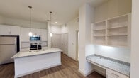 Located along W Highway 287 Access Road in Waxahachie, TX, this single-story unit offers a clean, modern aesthetic throughout. The open-concept kitchen serves as the heart of the space, showcasing white shaker-style cabinetry, granite countertops, a central island with an undermount sink, and a full suite of stainless steel appliances including a range, over-the-range microwave, and refrigerator. Sleek pendant lighting hangs above the island, adding a refined touch. Adjacent to the kitchen, a built-in desk nook with matching granite countertop and open shelving provides a dedicated workspace. The living area is generously proportioned, featuring warm hardwood-style flooring, a ceiling fan, and a window that invites natural light into the space. The bedroom is well-sized with its own ceiling fan, natural light from a window, and hardwood-style flooring that carries throughout. The en suite bathroom is a standout feature, offering a double-sink granite vanity with framed mirrors, marble-style tile flooring, and a glass-enclosed walk-in shower with elegant tile surround. A walk-in closet connects directly to the bathroom, completing the suite. The overall design palette of crisp whites and neutral tones creates a bright, cohesive, and move-in-ready environment.