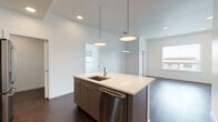 This single-floor residence showcases a clean, contemporary design throughout. The open-concept living and kitchen area serves as the heart of the home, anchored by a large central island with a white quartz countertop, an integrated sink, and a stainless steel dishwasher. Pendant lighting illuminates the island from above, while recessed lighting throughout ensures a bright and well-lit environment. The kitchen is fully equipped with stainless steel appliances, including a gas range, over-the-range microwave, and a French door refrigerator, all complemented by flat-front cabinetry in a warm dark tone. Rich, dark-stained hardwood flooring runs seamlessly through the main living areas, adding warmth and contrast to the crisp white walls. The living and dining areas open toward large windows and a glass-panel door leading to a private balcony, offering urban skyline views. The bedroom features plush neutral carpeting, expansive windows with roller shades, and direct balcony access that invites natural light. A well-organized reach-in closet with built-in shelving provides ample storage. The overall aesthetic is modern and minimalist, with a cohesive palette of whites, grays, and natural wood tones creating a refined and functional living environment.