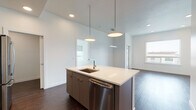This single-floor residence showcases a clean, contemporary design throughout. The open-concept living and kitchen area serves as the heart of the home, anchored by a large central island with a white quartz countertop, an integrated sink, and a stainless steel dishwasher. Pendant lighting illuminates the island from above, while recessed lighting throughout ensures a bright and well-lit environment. The kitchen is fully equipped with stainless steel appliances, including a gas range, over-the-range microwave, and a French door refrigerator, all complemented by flat-front cabinetry in a warm dark tone. Rich, dark-stained hardwood flooring runs seamlessly through the main living areas, adding warmth and contrast to the crisp white walls. The living and dining areas open toward large windows and a glass-panel door leading to a private balcony, offering urban skyline views. The bedroom features plush neutral carpeting, expansive windows with roller shades, and direct balcony access that invites natural light. A well-organized reach-in closet with built-in shelving provides ample storage. The overall aesthetic is modern and minimalist, with a cohesive palette of whites, grays, and natural wood tones creating a refined and functional living environment.