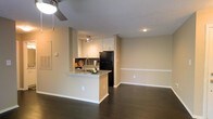 This single-floor residence offers an open-concept layout that seamlessly connects the kitchen and living areas, creating a spacious and cohesive environment. The kitchen is a standout feature, equipped with white cabinetry that extends to the ceiling for ample storage, a granite or stone-topped peninsula with a built-in sink, and a full-sized black refrigerator. A track lighting fixture above the kitchen ensures the workspace is well-lit, while recessed lighting adds to the overall brightness of the space. The peninsula doubles as a casual dining or prep area, adding both functionality and a social element to the kitchen. Rich, dark hardwood flooring runs throughout, providing a warm and elegant contrast to the neutral, beige-toned walls. A chair-rail detail along the walls adds a subtle architectural touch. The adjoining living area is open and well-proportioned, benefiting from a ceiling fan that promotes air circulation. The overall ambiance is clean, modern, and understated, with a neutral palette that offers a versatile backdrop for a variety of interior styles. The thoughtful design and quality finishes make this a practical and inviting space.