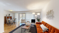 This thoughtfully designed single-floor residence showcases a cohesive mid-century modern aesthetic throughout. The open-concept living area is bright and welcoming, featuring warm parquet flooring, large sliding glass doors with vertical blinds, and generous natural light that highlights the clean white walls. A tufted orange sofa anchors the living space, complemented by a stylish arc floor lamp and a wood-and-metal coffee table, creating an inviting atmosphere.

Adjacent to the living area, the dining space accommodates a round walnut dining table with upholstered chairs, a wooden hutch, and direct access to a private balcony overlooking lush greenery. The kitchen is well-equipped with rich cherry wood cabinetry, granite countertops, a stainless steel gas range, dishwasher, and a tile backsplash, offering both function and style.

The bedroom is a serene retreat with plush carpeting, multiple windows allowing ample natural light, and access to outdoor views. A spacious walk-in closet with built-in shelving provides generous storage. The bathroom features a full soaking tub with clean white tile surround accented by a decorative mosaic tile border. The entry foyer includes a sleek console table, completing this well-curated and functional living space.
