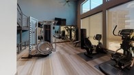 This fitness center is a thoughtfully designed, single-floor facility that offers a comprehensive workout environment. The space features soaring ceilings that contribute to an open, airy atmosphere, complemented by large, floor-to-ceiling windows dressed with roller shades. These windows allow ample natural light to fill the room while offering views of an outdoor pool area, creating an energizing backdrop for workouts.

The flooring throughout consists of striped, neutral-toned carpet tiles that add a modern aesthetic while providing comfort underfoot. The walls are finished in a cool blue-grey tone, lending the space a clean, contemporary feel.

The cardio zone is anchored by a row of ProMaxima treadmills positioned along the windowed wall, alongside an elliptical machine and a recumbent stationary bike. Wall-mounted flat-screen televisions are positioned for easy viewing from the cardio equipment. The strength training area includes a multi-functional cable machine, a weight rack stocked with dumbbells, a weight bench, and a mini trampoline. A large mirror extends along one wall, visually expanding the space and aiding in form monitoring. Recessed lighting and a ceiling fan provide additional comfort, while glass double doors offer easy access to the facility.