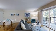 This well-appointed 1-bedroom, 1-bathroom residence is thoughtfully designed to maximize both comfort and functionality across a single floor. The living area serves as the heart of the space, showcasing warm hardwood flooring and smooth, clean-lined walls painted in a soft neutral tone that enhances the sense of openness and brightness. Large sliding glass doors dressed with flowing white curtains invite an abundance of natural light and provide access to an outdoor area, further expanding the sense of space. The living room is arranged to accommodate both relaxation and productivity, with a dedicated workspace area nestled alongside the main seating arrangement — a generous sectional sofa paired with a teal accent chair and a round coffee table, all set atop a patterned area rug that adds visual interest to the room. Vibrant abstract artwork adorns the walls, contributing a contemporary aesthetic throughout. A compact dining area with a round white table and modern chairs rounds out the living space, offering a clean and functional spot for everyday dining. The overall ambiance of this residence is modern, airy, and welcoming, making it an ideal space for comfortable everyday living.