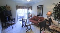 This single-floor residence offers a thoughtfully appointed living space that blends classic elegance with comfortable design. The living room serves as the heart of the home, featuring plush wall-to-wall carpeting and warm neutral tones that create an inviting atmosphere. Crown molding frames the ceiling, adding a refined architectural touch, while a ceiling fan provides practical comfort. A large sliding glass door floods the room with natural light and opens to a private balcony, where outdoor seating is visible — ideal for enjoying fresh air and outdoor views. Decorative window treatments with layered valances and drapes add a classic, tailored look to the windows. The living room is anchored by a richly carved wooden coffee table, complemented by a deep red upholstered sofa adorned with coordinating accent pillows. Tasteful artwork and decorative accessories are displayed throughout, contributing to the space's warm and curated character. A side console with decorative vases and greenery adds a natural element to the room. The overall ambiance is cozy and traditional, offering a welcoming environment that balances style and everyday livability.
