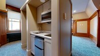 Located at 888 Delaware Ave in Buffalo, NY, this one-bedroom, one-bathroom unit blends historic architectural character with modern updates. The spacious bedroom is the centerpiece of the home, showcasing rich warm-toned wood wainscoting, ornate column details, and a striking decorative fireplace framed by an arched mural and vivid green ceramic tile surround — a true standout feature that speaks to the building's storied past. Plush blue-gray carpet runs throughout, complementing the warm woodwork. A cleverly integrated Murphy bed with gold hardware offers versatile use of the generous floor space, while a built-in wood cabinet provides additional storage and display options. Natural light filters in through multiple windows with classic wood framing. The compact kitchenette is efficiently tucked away, featuring white cabinetry, a marble-look countertop, an under-counter refrigerator, microwave, and a small sink — all the essentials neatly organized. The bathroom is finished to a high standard, with marble-look tile walls and flooring, a single vanity with a white countertop, a large mirror, and a glass-enclosed walk-in shower. Hallways connect the living spaces, contributing to a smooth and functional layout throughout this characterful unit.