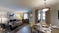 This well-appointed single-floor residence showcases a thoughtfully designed open-concept layout that seamlessly connects the living and dining areas, creating a spacious and cohesive atmosphere. The living area is anchored by a comfortable gray sofa, a sleek trunk-style coffee table, and tasteful wall art, including a striking equestrian print that adds a bold focal point to the room. Warm, neutral-toned walls complement the rich hardwood-style flooring, lending a sense of warmth and continuity throughout the space. A decorative sunburst mirror and a classic dresser add character and depth to the living area, while multiple windows dressed with elegant drapes invite generous amounts of natural light. The dining area flows naturally from the living space and features a round table with woven accent chairs, set beneath a contemporary pendant light fixture. Patterned drapery panels frame the windows, adding visual interest and a refined touch. The overall ambiance strikes a balance between casual comfort and sophisticated style, making the space feel both inviting and polished. The open sightlines and cohesive design elements throughout contribute to an airy, well-proportioned interior that is ideal for both everyday living and entertaining.
