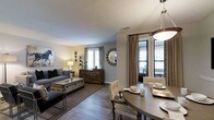 This well-appointed single-floor residence showcases a thoughtfully designed open-concept layout that seamlessly connects the living and dining areas, creating a spacious and cohesive atmosphere. The living area is anchored by a comfortable gray sofa, a sleek trunk-style coffee table, and tasteful wall art, including a striking equestrian print that adds a bold focal point to the room. Warm, neutral-toned walls complement the rich hardwood-style flooring, lending a sense of warmth and continuity throughout the space. A decorative sunburst mirror and a classic dresser add character and depth to the living area, while multiple windows dressed with elegant drapes invite generous amounts of natural light. The dining area flows naturally from the living space and features a round table with woven accent chairs, set beneath a contemporary pendant light fixture. Patterned drapery panels frame the windows, adding visual interest and a refined touch. The overall ambiance strikes a balance between casual comfort and sophisticated style, making the space feel both inviting and polished. The open sightlines and cohesive design elements throughout contribute to an airy, well-proportioned interior that is ideal for both everyday living and entertaining.