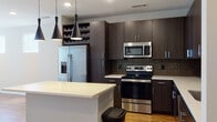 This single-floor unit showcases a contemporary design with a cohesive palette of dark espresso cabinetry, light countertops, and warm wood-style flooring throughout the main living areas. The kitchen is a standout feature, equipped with full stainless steel appliances including a French door refrigerator, electric range, and built-in microwave. A decorative mosaic tile backsplash adds visual texture behind the cooking area, while floor-to-ceiling dark cabinetry maximizes storage capacity. A built-in wine rack provides an elegant organizational detail. The central island, topped with a crisp light quartz surface, offers additional prep space and casual seating. Three sleek pendant lights hang above, providing focused task lighting with a modern aesthetic. The living area features warm hardwood-style flooring, multiple windows with blinds allowing natural light, and a ceiling fan for climate control. A secondary carpeted room with its own ceiling fan and windows serves well as a bedroom or flex space. The bathroom is notably spacious, featuring a lengthy dual-sink vanity with dark cabinetry and white countertops, large-format dark tile flooring, a separate glass-enclosed shower with tile surround, a private water closet, and a walk-in closet with built-in shelving.