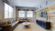 Situated at 100 Underwood Dr in Summerville, South Carolina, this single-floor space combines comfort and style through a carefully curated interior design. The main lounge area serves as the centerpiece of the space, featuring a generous leather sectional sofa paired with a round wooden coffee table that anchors the room with a warm, organic feel. Natural light pours in through multiple large windows dressed with classic white plantation shutters and decorative Roman shades in a bold geometric pattern, creating a bright and inviting atmosphere. A striking reclaimed wood accent wall adds rustic texture and visual depth, providing a natural backdrop for the wall-mounted flat-screen display and the stylish dark media console below. Crown molding, wainscoting, and recessed lighting throughout contribute a refined, polished character to the space. The neutral color palette of soft grays and warm taupes, complemented by the natural wood tones, creates a cohesive and welcoming ambiance. The overall design strikes a balance between casual comfort and elevated aesthetics, making this a functional and visually appealing gathering space.