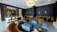 This single-floor space showcases a carefully curated interior design that blends contemporary elegance with a moody, upscale aesthetic. The lounge area is anchored by deep navy blue wall paneling accented with warm gold wall sconces, creating a rich and dramatic backdrop. A statement chandelier with layered gold rings serves as a striking focal point overhead, complementing the recessed lighting throughout the space.

The seating arrangement centers around a curved caramel-toned sofa that defines the main lounge zone, paired with plush deep purple accent chairs adorned with chartreuse yellow throw pillows — a bold color combination that adds visual energy. Geometric stone-finished cube coffee tables ground the seating area, which sits atop a teal-toned area rug inset within the flooring.

Adjacent to the lounge, a bar or kitchenette area is visible, featuring dark cabinetry, pendant lighting in brushed gold finishes, and bar-height seating, suggesting a hospitality-oriented function. The overall space communicates a refined, boutique-style atmosphere well-suited for social gatherings or premium communal use.