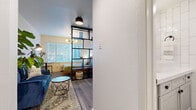 This well-appointed single-floor studio makes smart use of its layout through clever design choices that define distinct living zones without sacrificing openness. The main living area features light wood-tone flooring throughout and is anchored by a striking black open-grid shelving unit that elegantly separates the sleeping area from the lounge space while maintaining visual flow and allowing natural light to pass freely. The living zone accommodates a seating area with a patterned area rug, a geometric coffee table, and a vibrant blue sofa, all bathed in natural light from large windows fitted with horizontal blinds. The sleeping nook, nestled behind the room divider, offers a cozy, semi-private retreat with a window that brings in additional daylight. The compact kitchenette is efficiently appointed with gray shaker-style cabinetry, a light countertop, a single-basin sink with a matte black faucet, a decorative patterned tile backsplash, and under-counter refrigeration. A hallway leads to the entry door, keeping the main living space unencumbered. The bathroom is finished with floor-to-ceiling vertically stacked white tile walls, dark hexagonal floor tiles, a white vanity with matte black hardware, and a curtained shower area, creating a clean, modern aesthetic throughout.