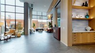 This contemporary lobby space makes an immediate impression with its soaring double-height ceilings and expansive floor-to-ceiling glazing that floods the interior with natural light while framing views of the surrounding streetscape and outdoor courtyard. The signature hexagonal grey tile flooring flows seamlessly throughout, anchoring the open, airy layout and lending a refined geometric character to the space.

The lobby is organized into several distinct yet connected zones. A welcoming reception area features a rich wood-paneled desk accented by warm natural wood columns and architectural detailing. Adjacent lounge seating areas are thoughtfully furnished with mid-century modern chairs and curated greenery, including statement fiddle-leaf fig trees. One lounge zone is anchored by a vibrant large-scale abstract mural in bold teal tones, serving as a dramatic focal point alongside a sleek slatted wood partition wall.

A fully equipped catering kitchen offers light wood cabinetry with white countertops, a large central island, grey subway tile backsplash, open display shelving, and integrated appliances — ideal for events and gatherings. Toward the rear, another seating area opens directly to outdoor pool and courtyard views through a second dramatic glass wall. Throughout, recessed and pendant lighting complement the warm wood tones, creating a polished and inviting ambiance.