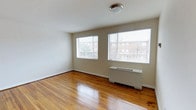 This single-floor space offers a clean and functional layout with a welcoming, light-filled ambiance. The main living area is immediately notable for its richly toned, polished hardwood floors that stretch across the room, reflecting natural light and adding warmth to the overall atmosphere. Two generously sized windows fitted with horizontal blinds line one wall, offering views of the surrounding neighborhood and allowing ample daylight to fill the space. The walls are finished in a soft, neutral tone that complements the flooring and contributes to the room's open, airy feel. A wall-mounted heating unit sits beneath the windows, seamlessly integrated into the room without interrupting the clean lines of the space. Overhead, a simple flush-mount ceiling light provides even illumination throughout. The room's straightforward design and well-maintained finishes make it a versatile space adaptable to a variety of uses. White baseboards frame the perimeter of the room, adding a crisp finishing touch that ties together the interior palette. Overall, the space presents a tidy, move-in-ready environment with classic architectural details and a practical, uncluttered character.
